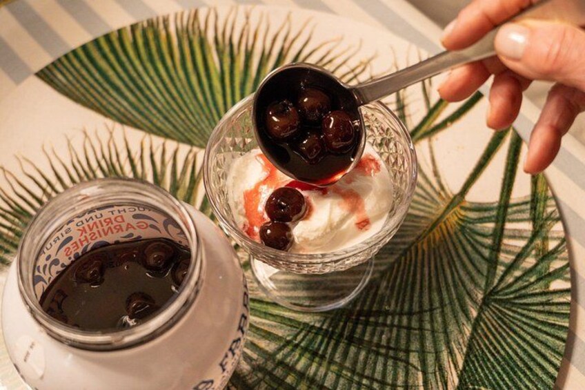 Un tazón de yogur con cerezas y una cuchara, un tarro de líquido oscuro y una hoja de palma. 3