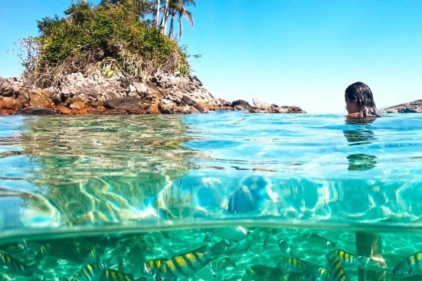 Helder turquoise water, een rotsachtig eiland met begroeiing, en iemand die zwemt. 1