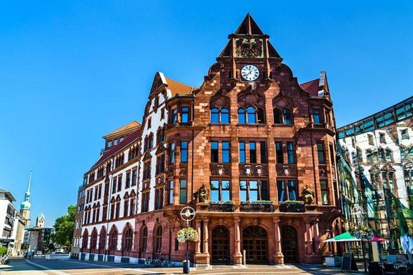 A historic red brick building with a clock tower, arched windows, and a balcony with hanging plants. 2