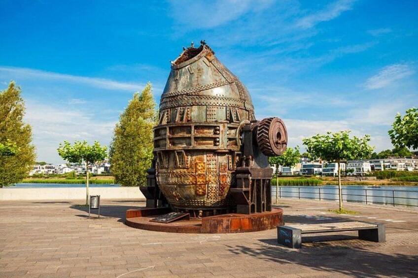 A large, rusted, spherical structure with a cylindrical base, surrounded by a paved area with benches. 3