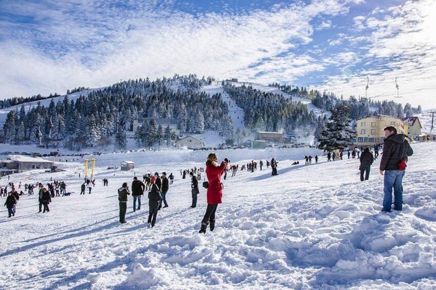 A snowy landscape with people walking and standing, a ski lift, and a building in the background. 1