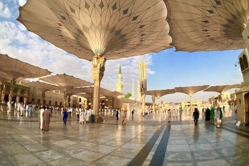 A large open courtyard with people walking, covered by large, ornate umbrellas, and a tall minaret in the background. 1