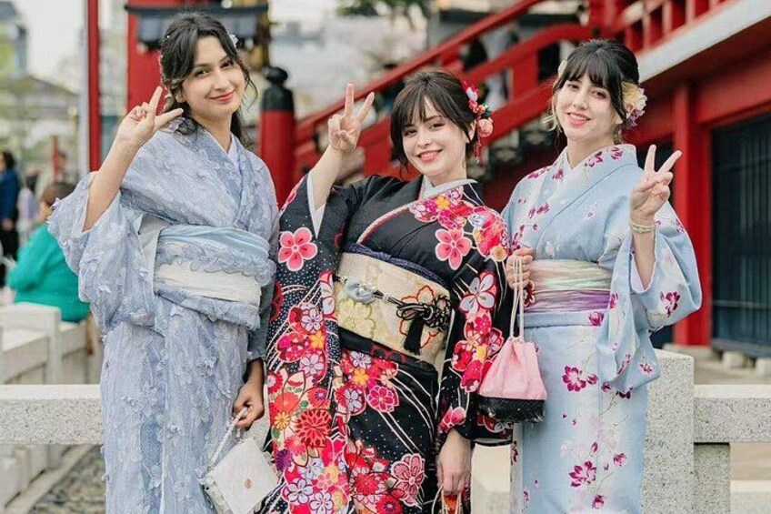 Drie mensen in traditionele Japanse kimono's met bloemenmotieven, voor een rode gebouw met een pad van stenen. 2