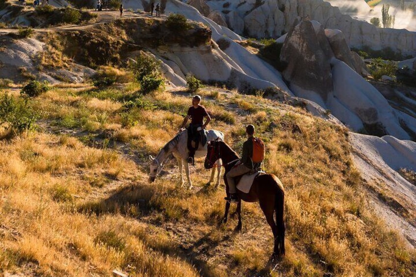 Zwei Personen reiten auf einer grasbewachsenen Anhöhe, im Hintergrund Felsformationen. 3
