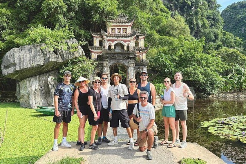 A group of people standing in front of a traditional temple with a pond and greenery in the background. 1