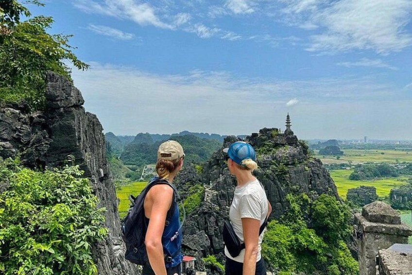 Dos personas están sobre un promontorio rocoso, contemplando un valle con vegetación y una pagoda al fondo. 5