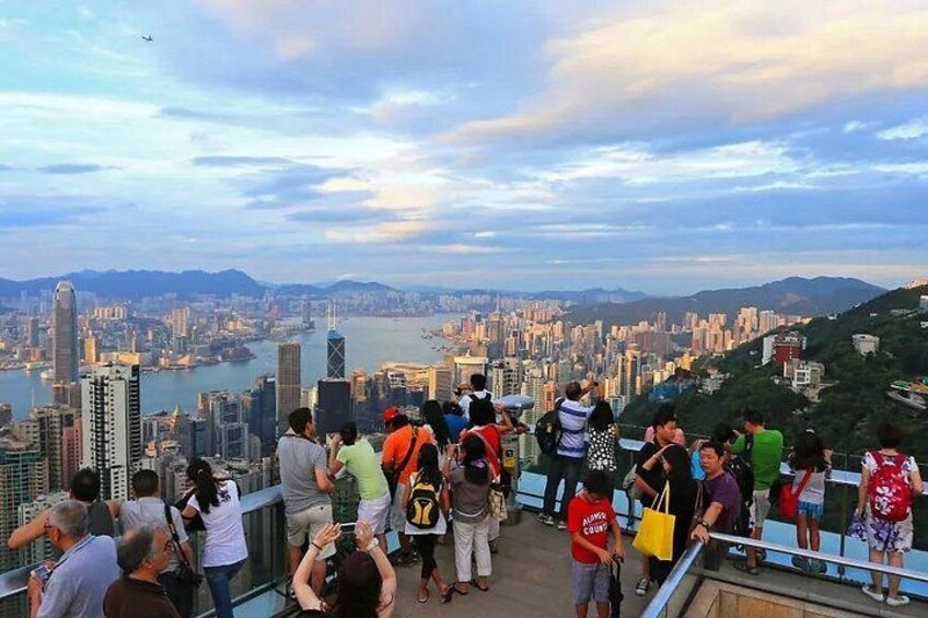 A crowded viewpoint overlooking a cityscape with a river, mountains, and a clear sky. 4