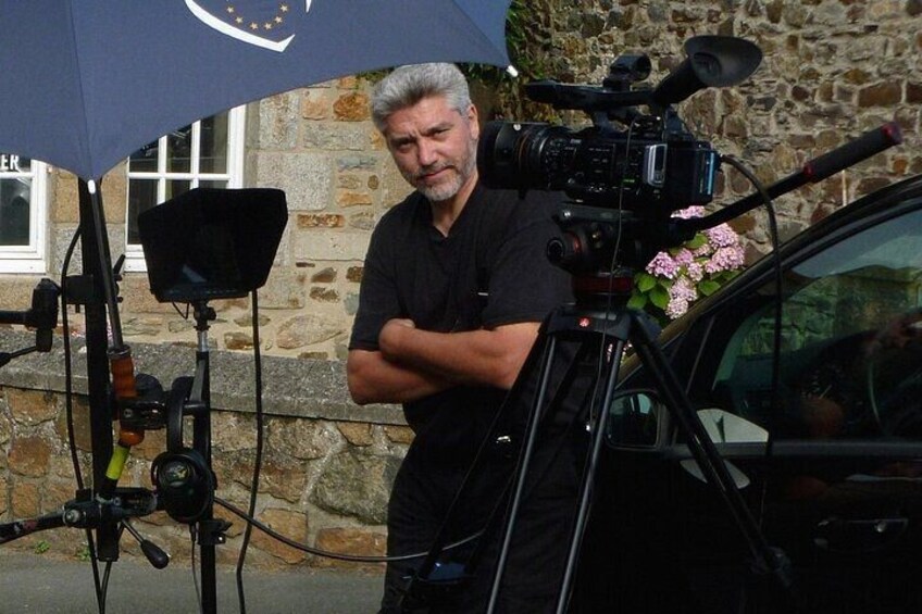 A man standing next to a camera on a tripod with a car in the background. 2