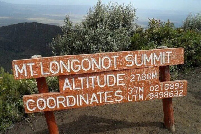 Un cartello di legno con il testo 'Altitudine della vetta del Monte Longonot 2780M' e coordinate '37M 0215892 UTM 9898632'. 3