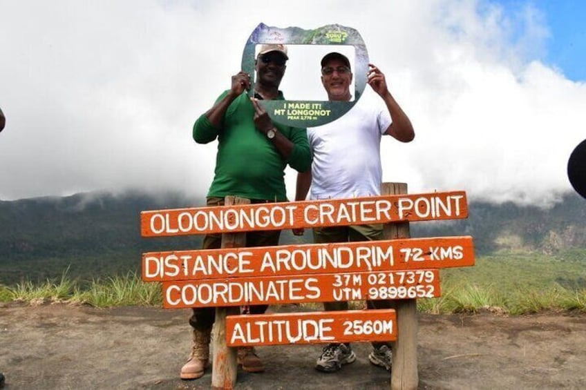 Due persone che reggono un cartello con informazioni su Oloonongot Crater Point, tra cui la distanza del perimetro, le coordinate e l'altitudine. 4