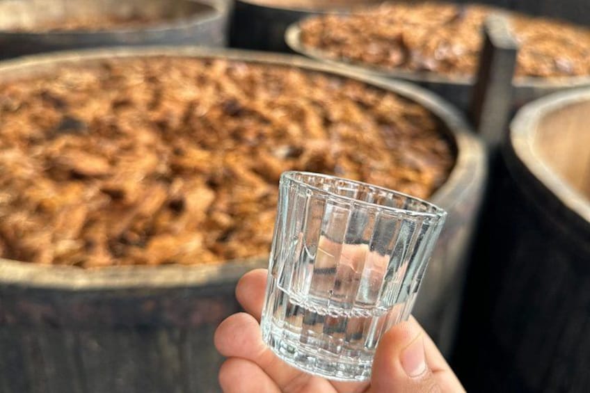 A hand holding a clear glass with a liquid, in front of large containers filled with a brownish substance. 5