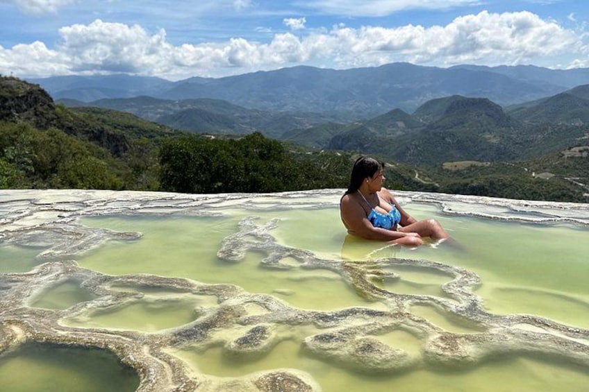 A person sitting in a natural hot spring surrounded by green hills and mountains. 1