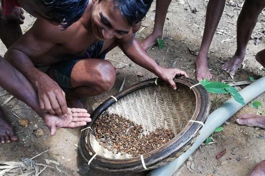 A person handling a woven basket filled with seeds, standing on a dirt ground with a green pipe nearby. 5