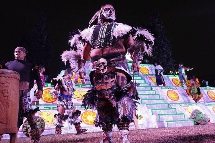 Un evento serale all'aperto con artisti in costumi elaborati, una struttura a piramide e uno sfondo con motivi di frutta e forme geometriche. 5