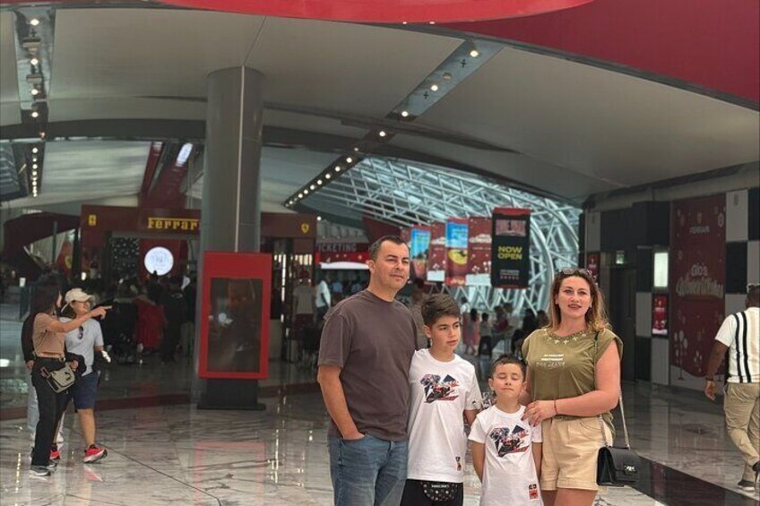 Une famille de cinq personnes pose dans un centre commercial moderne, à l’intérieur, où l’on peut voir des panneaux publicitaires pour Ferrari et d’autres marques. 4