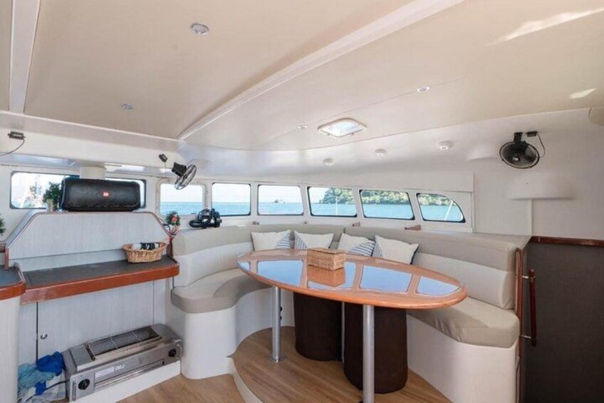 A boat interior with a dining area, a small kitchen, and a seating area with windows. 5