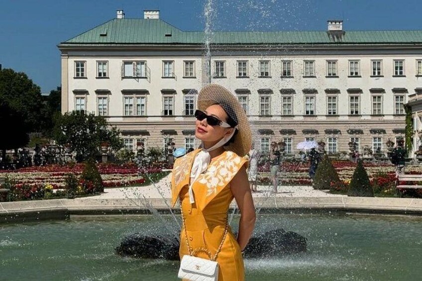 A person in a yellow dress standing in front of a fountain with a large building in the background. 1