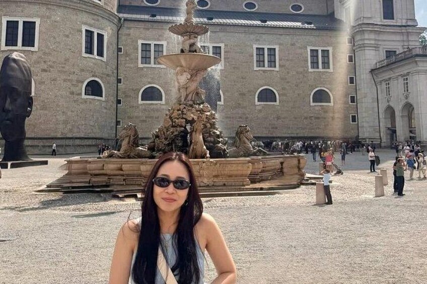 A woman in front of a fountain with sculptures and a large building in the background. 4