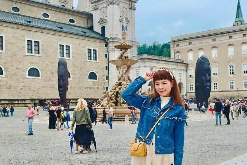 A person standing in a public square with a fountain and large buildings in the background. 3