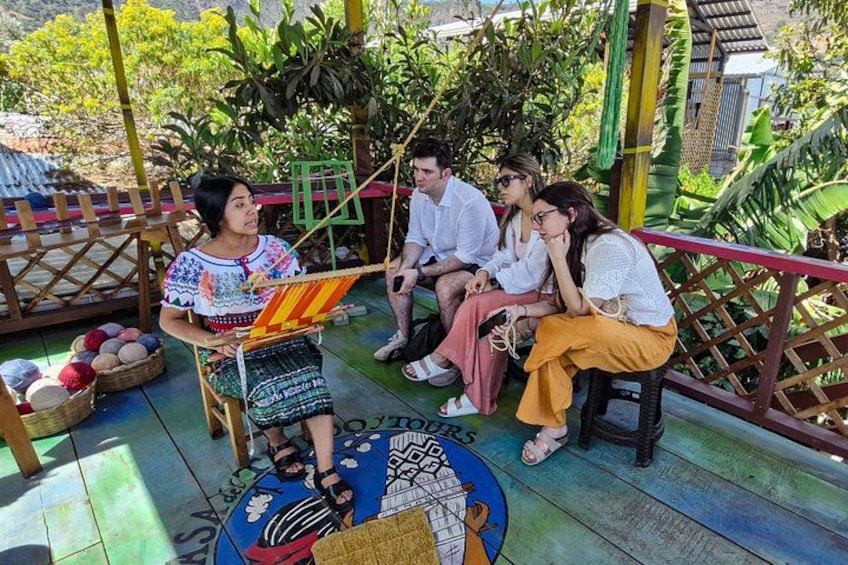 A group of people sitting on a wooden deck with a colorful woven mat, surrounded by lush greenery and a wooden fence. 4