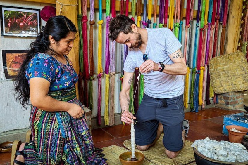 A person kneeling on a mat, working with a long white object, possibly a tool or utensil, in a room with colorful hanging items and a woven basket in the background. 5
