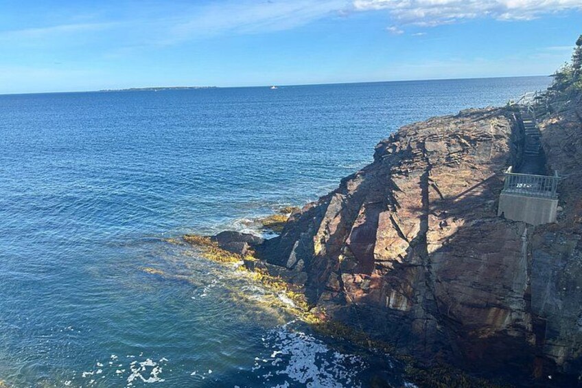 Paisaje costero con acantilados rocosos, una escalera que baja hacia el mar y un cielo azul despejado. 4