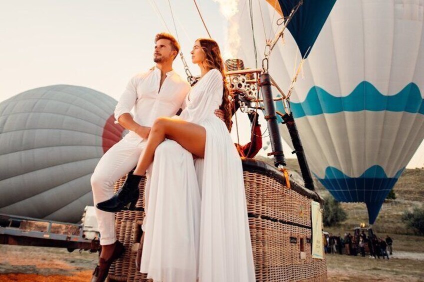 A couple on a hot air balloon with a wicker basket, a large white balloon with blue waves, and a group of people on the ground. 2