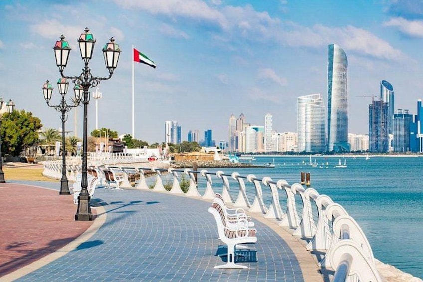 A waterfront promenade with a row of white benches, a candelabra street lamp, and a city skyline in the background. 2