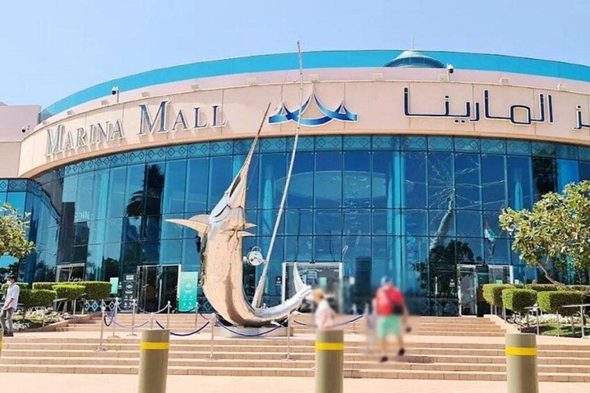 A modern building with a large glass facade, a prominent sailboat sculpture, and Arabic text. 3