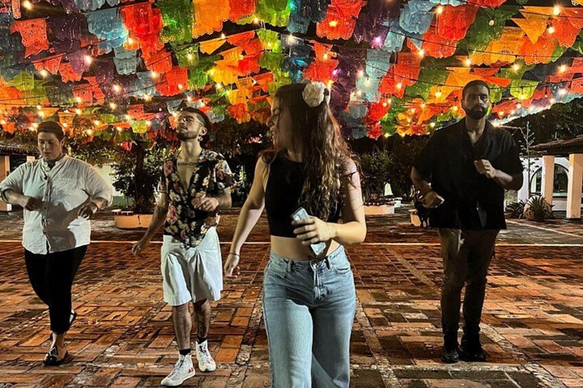 Personas bailando en una plaza con coloridas decoraciones de papel colgando. 1