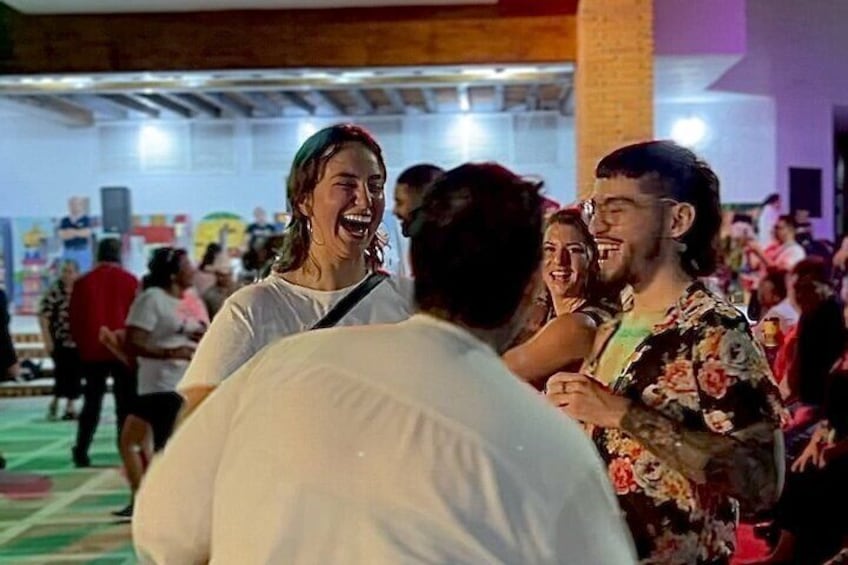 Un gruppo di persone a un evento sociale, forse una festa, in un ambiente interno con un muro di mattoni e decorazioni colorate sullo sfondo. 3