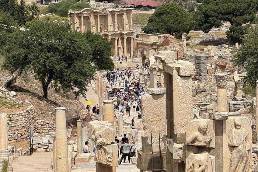 Ancient ruins with columns and statues, a crowd of visitors, and a large building in the background. 1