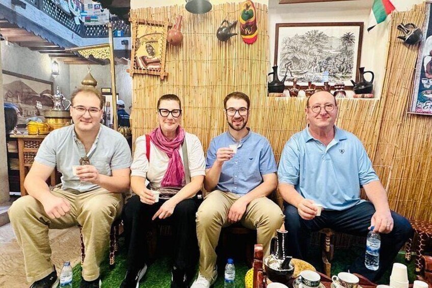 Quatre personnes sont assises dans une pièce aux murs en bambou, entourées de divers objets et d’une table sur laquelle se trouvent des tasses et des bouteilles. 3