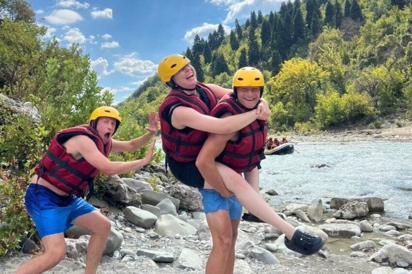 Three people in life jackets and helmets near a river with rocks and trees in the background. 2
