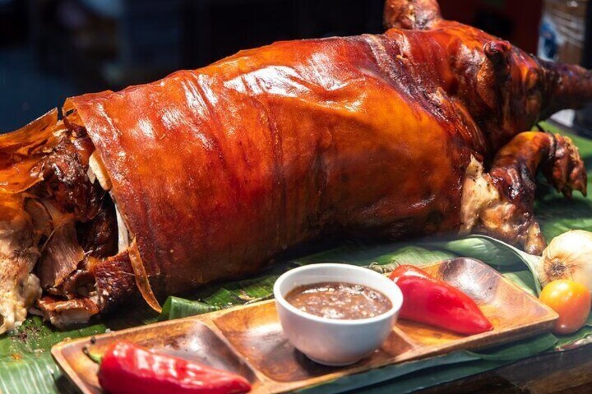 A roasted pig on a banana leaf with a bowl of sauce and sliced red peppers. 1