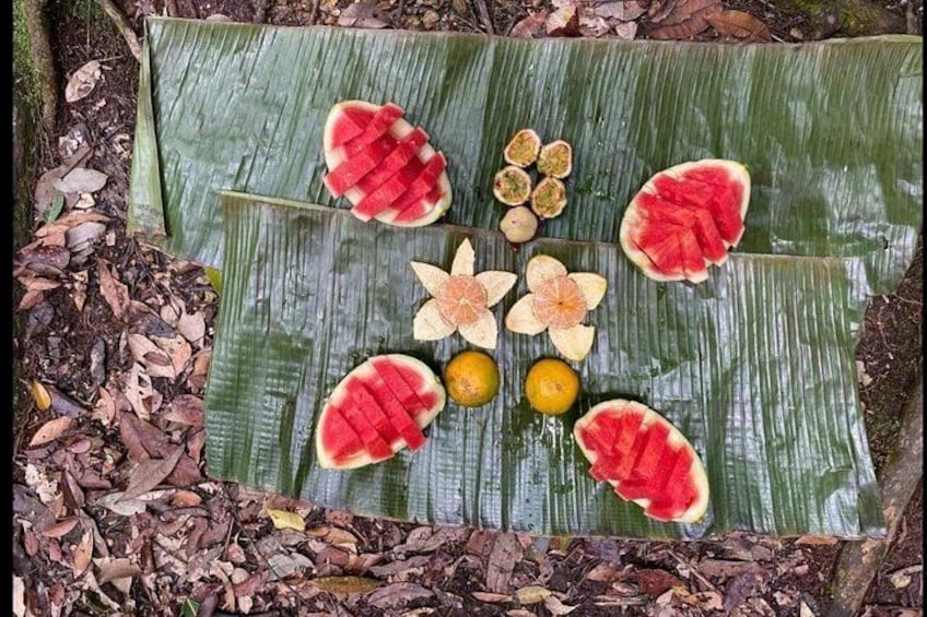 Una hoja de plátano con sandía en rodajas, cítricos y pétalos de flores dispuestos sobre el suelo. 2
