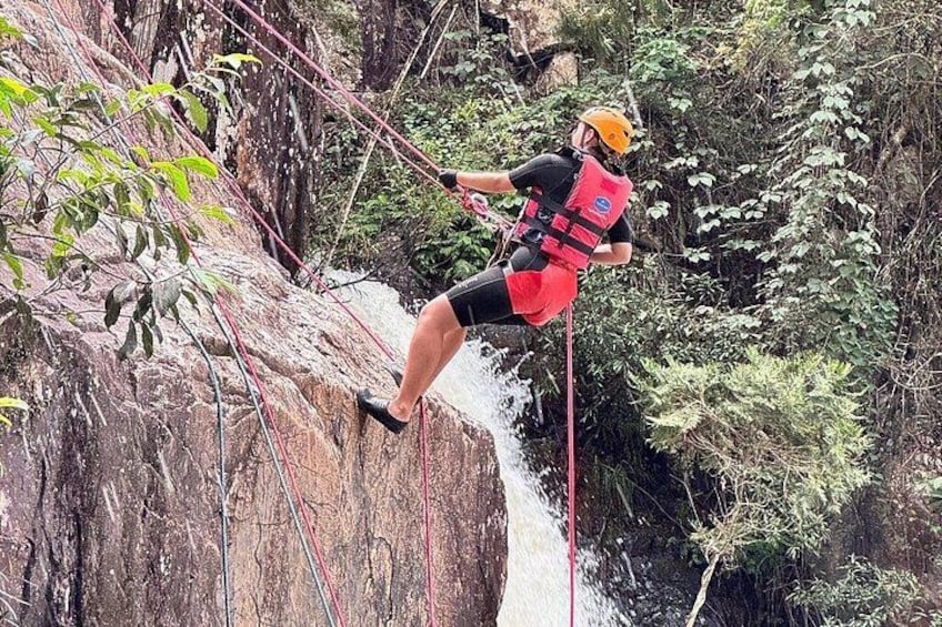 A person rappelling down a rocky cliff with a harness and helmet, surrounded by dense forest. 5