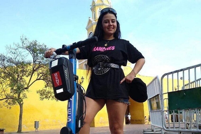 En person står ved siden af en blå scooter med en rød etiket, iført en sort t-shirt med lyserød tekst og jeansshorts. 4