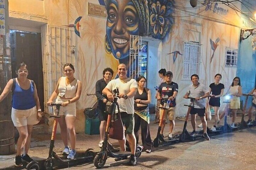A group of people riding electric scooters on a street with murals on the walls. 1