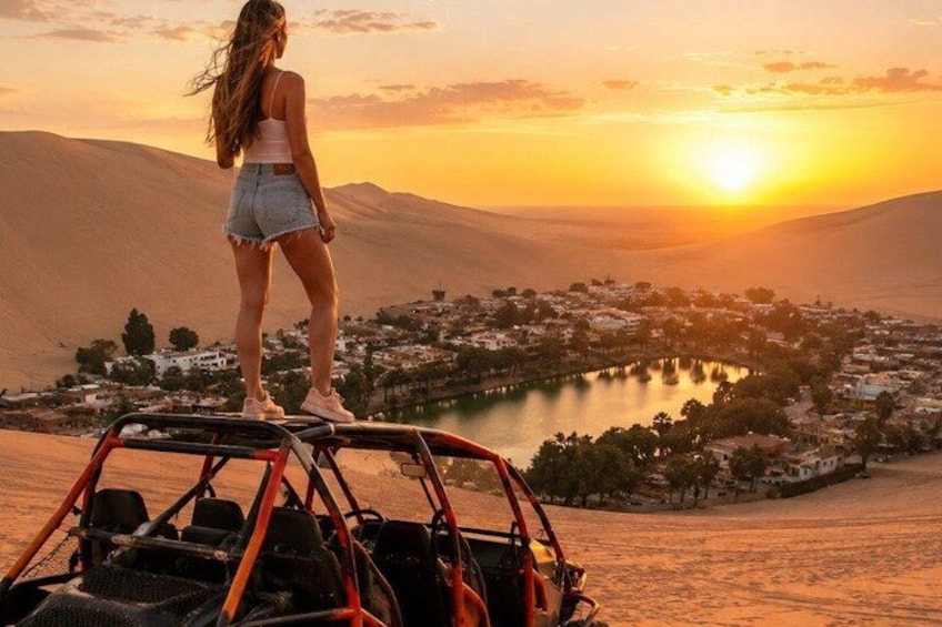 A person standing on a dune with a view of a town and a lake at sunset. 1