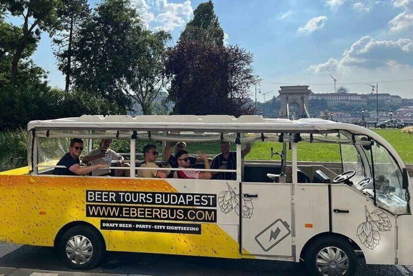 A yellow and white bus with passengers, labeled "Beer Tours Budapest," parked outdoors with trees and a monument in the background. 1