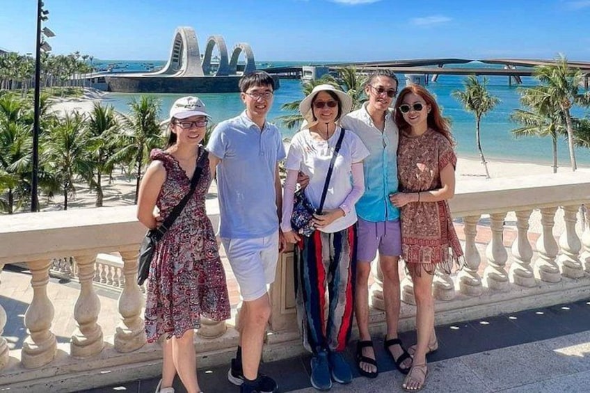 A group of people standing on a balcony overlooking a beach with palm trees and a distinctive bridge structure in the background. 1