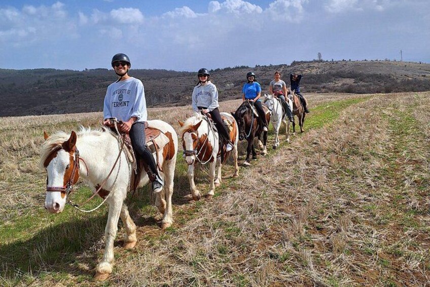 Eine Gruppe von Menschen reitet auf Pferden über eine Wiese, im Hintergrund eine hügelige Landschaft. 3