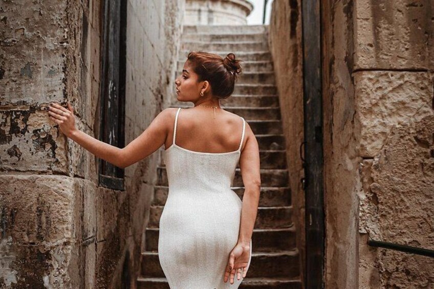 Una persona con un vestido blanco parada en un callejón con paredes de piedra y una escalera. 5