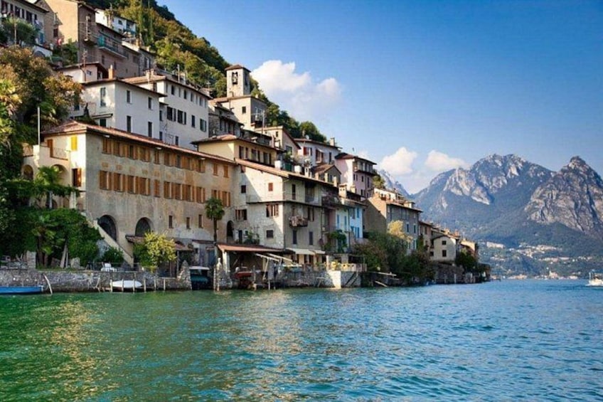 A coastal town with buildings on a hillside, a body of water, and mountains in the background. 1