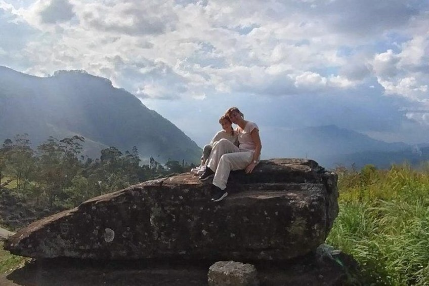 Two people sitting on a rock with a mountainous landscape in the background. 2