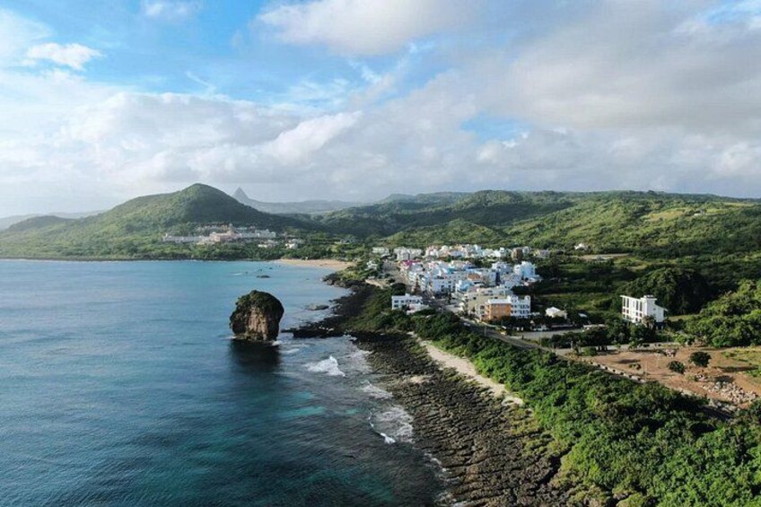A coastal town with buildings, a prominent rock formation, and green hills in the background. 1