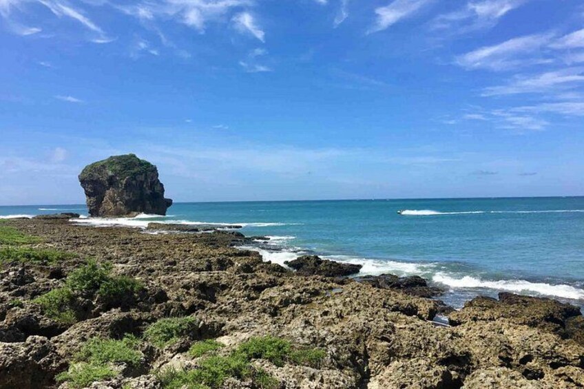A rocky coastline with a prominent rock formation in the sea, waves crashing against the shore, and a clear blue sky. 3