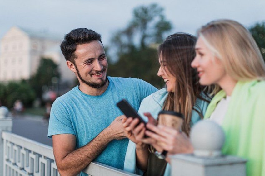 Tre personer står ved et gelænder, hvoraf den ene holder en smartphone og en anden en kaffekop. 4