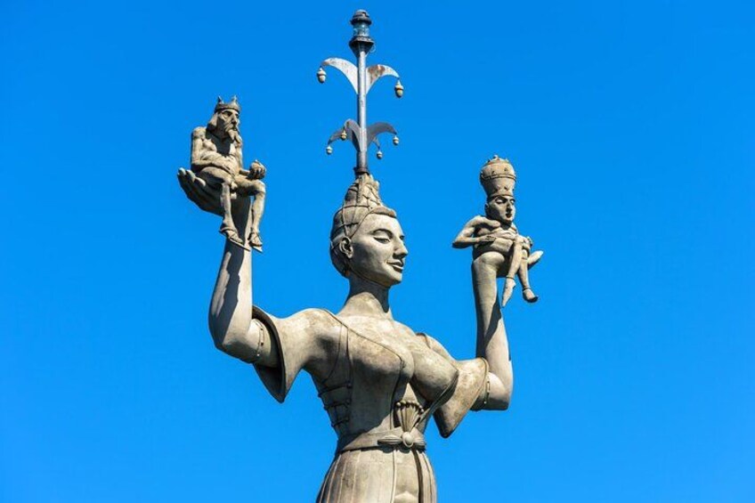 En staty som föreställer en kvinna som håller två figurer, en i varje arm, och som har ett kors på huvudet. 4